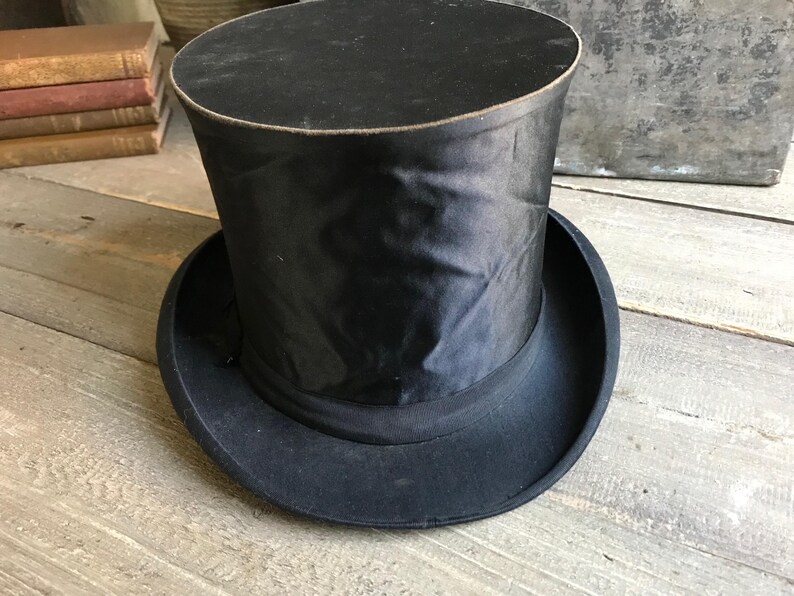 May include: A black top hat with a tall, cylindrical crown and a wide brim. The hat is made of a smooth, black fabric.