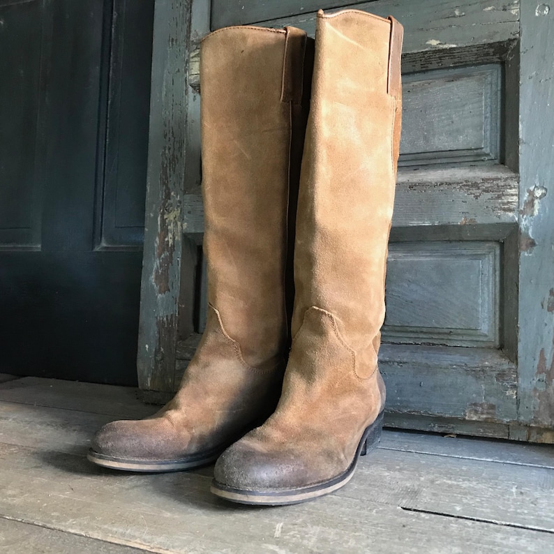 May include: A pair of tall, brown suede boots. The boots have a worn, distressed appearance with a slight heel and a classic, knee-high design. The boots are standing on a wooden surface against a weathered door.