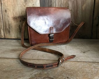 Pode incluir: Uma bolsa de couro marrom transversal com fecho de aba e fivela prateada. A bolsa tem uma alça longa ajustável.