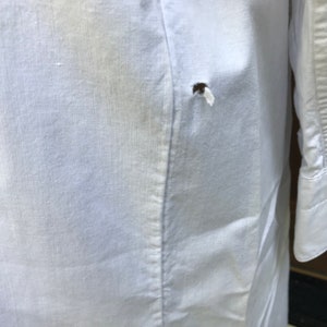 May include: White linen shirt with a small hole in the back. The shirt has a long sleeve and a button-up closure.