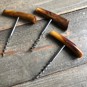 May include: Three vintage corkscrews with brown and yellow handles and silver metal corkscrews. The handles have a marbled appearance. The corkscrews are arranged on a weathered wooden surface.