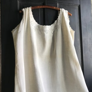May include: A cream-coloured vintage slip dress with a delicate floral lace trim along the neckline. The sleeveless dress hangs from a wooden hanger against a dark background. The fabric appears to be lightweight and slightly textured.