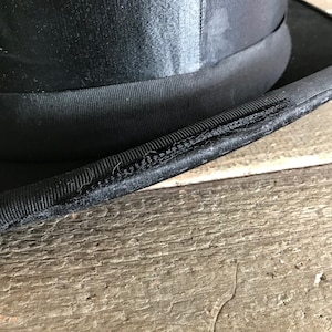 May include: Close-up of the black fabric brim of a top hat. The brim is stitched together with a visible seam.