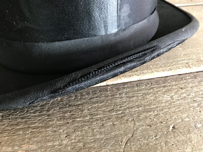 May include: Close-up of the black fabric brim of a top hat. The brim is stitched together with a visible seam.