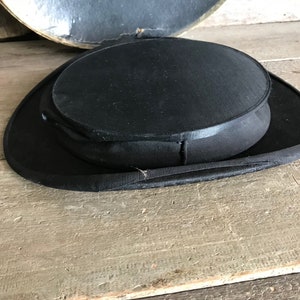 May include: A black top hat with a wide brim and a tall, round crown. The hat is made of a smooth, felt-like material.