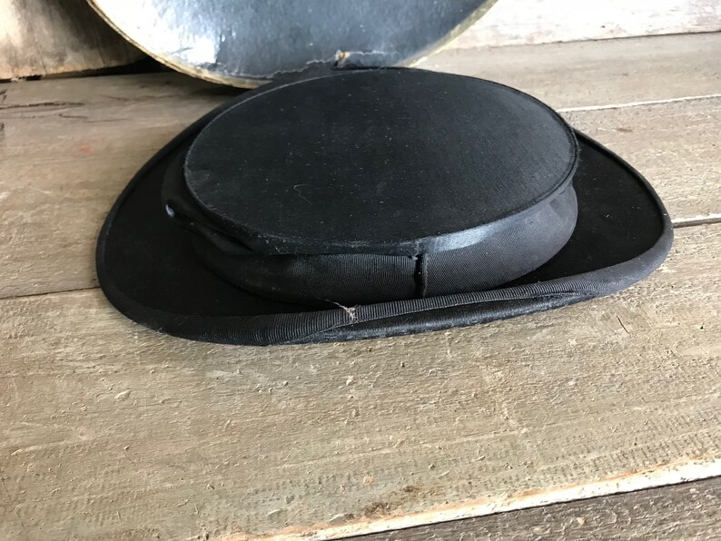 May include: A black top hat with a wide brim and a tall, round crown. The hat is made of a smooth, felt-like material.