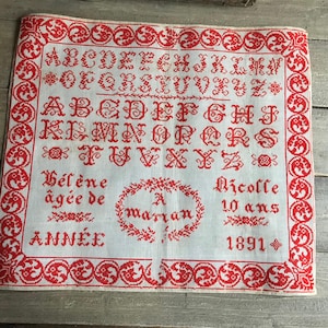 以下が含まれることがあります： 白地に赤い刺繍が施されたヴィンテージのクロスステッチのサンプラー。 サンプラーには、アルファベット、装飾的なボーダー、名前、年齢、1891年などのテキストが記載されています。 デザインは手仕事の古典的な例です。