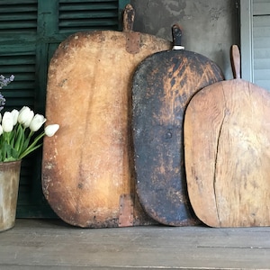 Pode incluir: Três tábuas de pão de madeira vintage de tamanhos diferentes. As tábuas são de forma oval e têm um aspeto rústico e desgastado. A tábua maior é de cor castanho claro, a tábua do meio é de cor castanho escuro e a tábua mais pequena é de cor castanho médio.