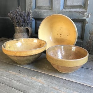 May include: Three antique ceramic bowls in varying sizes, featuring a warm yellow and brown color palette. The bowls have a rustic, aged appearance, with a wide rim and a speckled interior. A small, textured, brown ceramic pitcher with lavender is in the background.