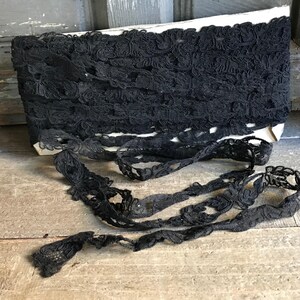 Puede incluir: Un rollo de encaje negro con un diseño floral. El encaje se muestra sobre una base de cartón blanco, con parte del encaje desenrollado sobre una superficie de madera. El encaje presenta un delicado patrón calado.