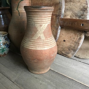 May include: A brown clay jug with white painted lines and an X design. The jug is sitting on a wooden surface.