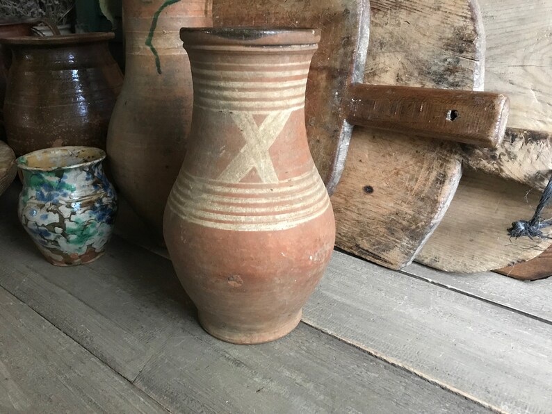 May include: A brown clay jug with white painted lines and an X design. The jug is sitting on a wooden surface.