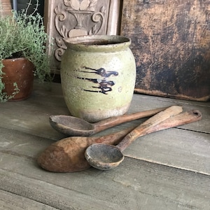 May include: A rustic still life featuring a light green ceramic jar with dark brown markings and three wooden spoons. The spoons vary in size and show signs of wear. The scene evokes a vintage, farmhouse aesthetic.