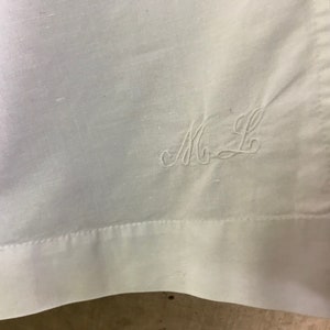 Pu&ograve; includere: Primo piano di un tessuto bianco con le iniziali ricamate "M L" in una scrittura delicata. Il tessuto ha una trama sottile e un bordo cucito. L'estetica generale &egrave; semplice ed elegante.