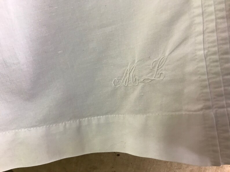 Pu&ograve; includere: Primo piano di un tessuto bianco con le iniziali ricamate "M L" in una scrittura delicata. Il tessuto ha una trama sottile e un bordo cucito. L'estetica generale &egrave; semplice ed elegante.
