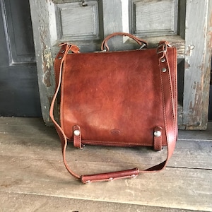 May include: A brown leather satchel with a top handle and adjustable shoulder strap. The bag has a rectangular shape with a flap closure and metal hardware. The leather has a distressed appearance.