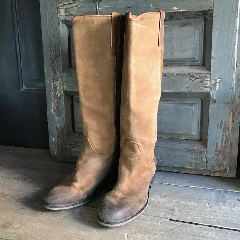 May include: A pair of knee-high, tan suede boots. The boots have a classic Western style with a pointed toe and a stacked heel. The boots are worn and have a vintage look, with some wear and tear visible on the suede.