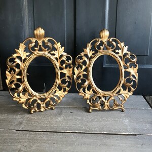 May include: Two ornate gold-coloured metal picture frames with intricate scrollwork and a leaf design. The frames are oval shaped and have a vintage look.