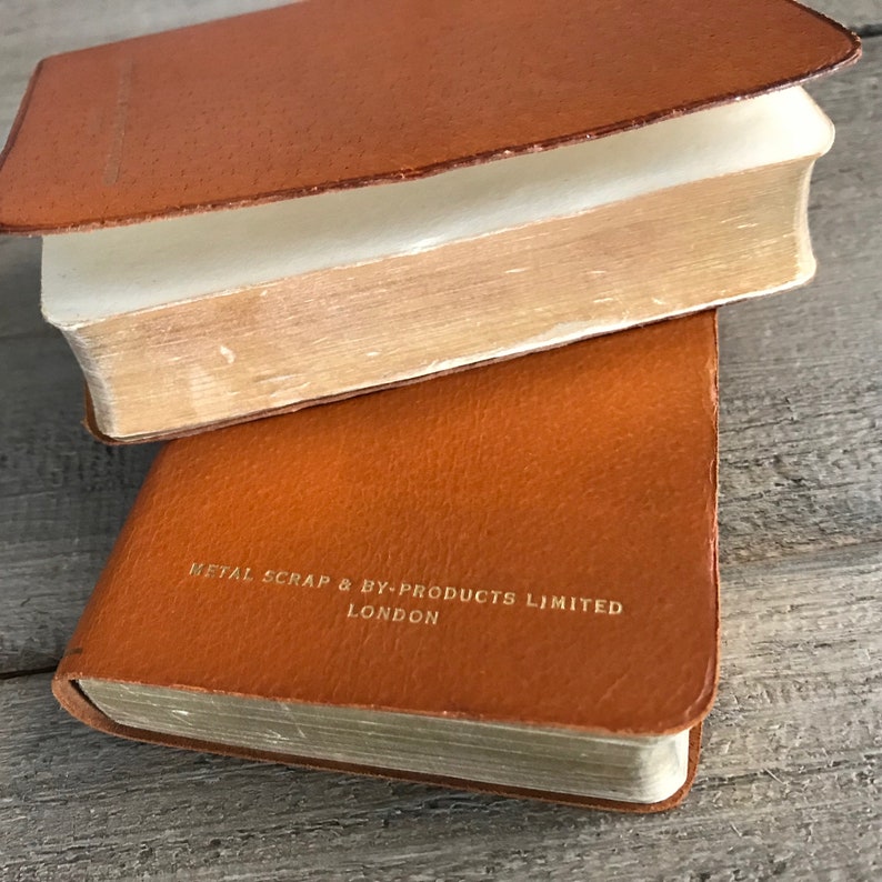 May include: Two brown leather-bound books stacked on top of each other. The top book is closed and the bottom book is open, revealing the pages. The bottom book has the text "METAL SCRAP & BY-PRODUCTS LIMITED LONDON" printed in gold on the front cover.