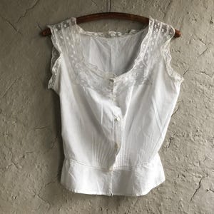 Puede incluir: Un camisón de algodón blanco con un ribete de encaje alrededor del escote y las sisas. El camisón tiene un cierre de botones en la parte delantera y un dobladillo con volantes.