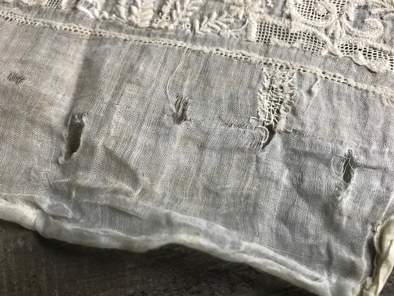 以下が含まれることがあります： 白い布のクローズアップ。いくつかの穴と繊細な刺繍模様があります。布は古びていて、ビンテージな外観をしています。