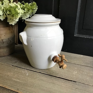May include: White ceramic crock with a wooden tap and a lid. The crock is on a wooden surface.