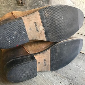 May include: Pair of worn brown suede boots with black rubber soles. The soles are marked with the brand name "BRONX" and size "39". The boots show signs of wear and tear, suggesting they are well-used.