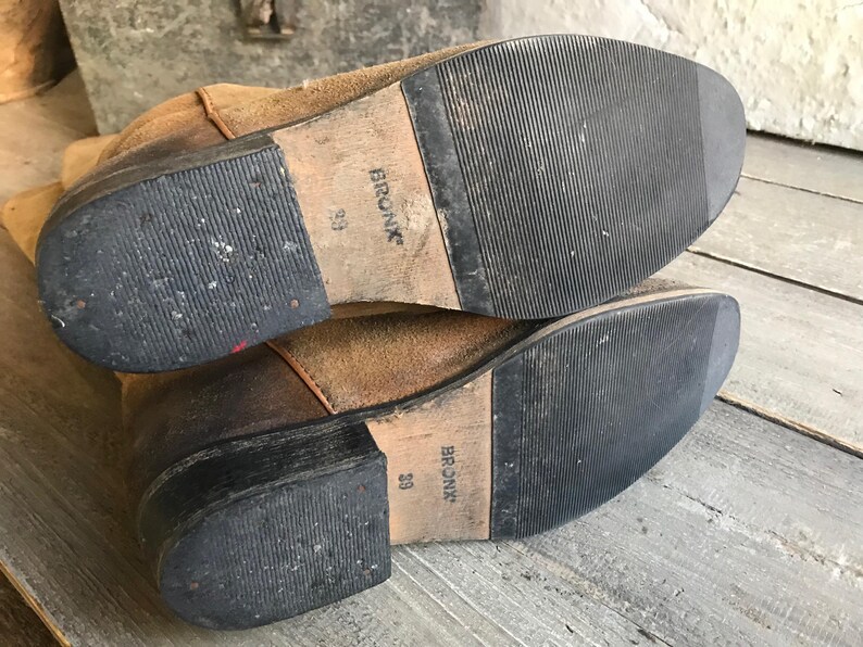 May include: Pair of worn brown suede boots with black rubber soles. The soles are marked with the brand name "BRONX" and size "39". The boots show signs of wear and tear, suggesting they are well-used.