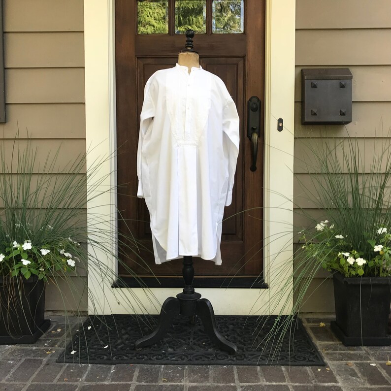 May include: A white long-sleeved shirt with a stand-up collar and a button-up front. The shirt is displayed on a black mannequin stand in front of a wooden door.