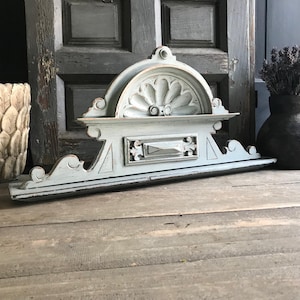 Può includere: Un elemento architettonico in legno azzurro chiaro e usurato. L'elemento decorativo presenta una parte superiore semicircolare con un motivo floreale e un pannello rettangolare sottostante. Il pezzo è invecchiato, con segni di usura visibili, e poggia su una superficie in legno.