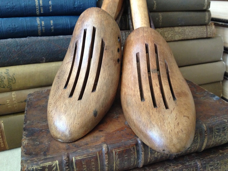 May include: Two wooden shoe trees with slotted tops, resting on antique books. The shoe forms are light brown with a smooth finish. The background features stacks of old books with various aged covers, creating a vintage aesthetic.