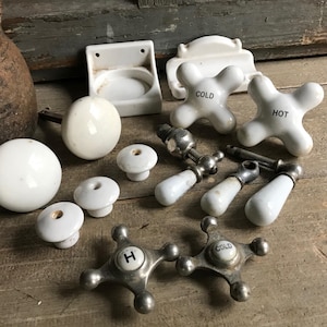 May include: A selection of antique white porcelain and metal bathroom fittings, including tap handles, knobs, and a soap dish. The tap handles are marked "COLD" and "HOT". The items are displayed on a weathered wooden surface.
