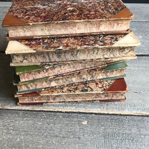 May include: A stack of vintage books with marbled covers in shades of brown, green, and cream. The books are arranged in a staggered pattern, with the top book slightly offset from the others.