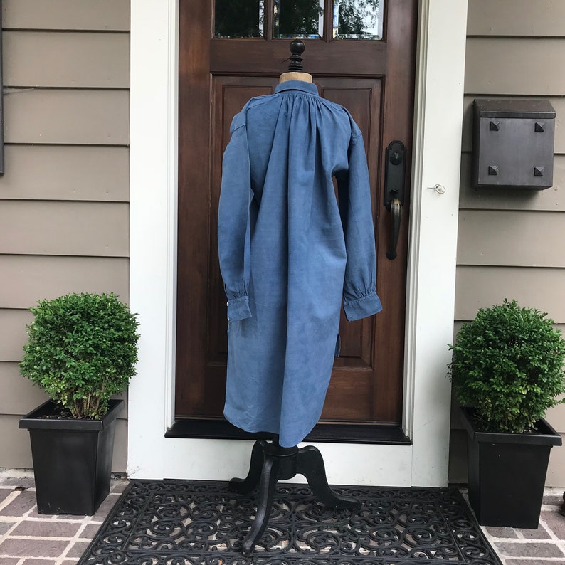 May include: A long-sleeved, blue denim shirt with a gathered neckline and long sleeves. The shirt is displayed on a mannequin against a wooden door.