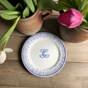 May include: White ceramic plate featuring a blue floral border and a central blue letter "C" in an ornate design. The plate is set amongst rustic pottery and fresh tulips, creating a vintage aesthetic.