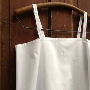 Puede incluir: Top blanco sin mangas de algodón con tirantes finos colgado de una percha de madera sobre un fondo de madera marrón.