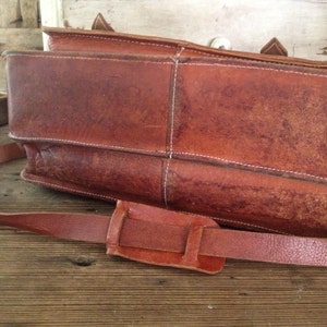 K&ouml;nnte beinhalten: Eine braune Ledertasche mit einem langen verstellbaren Riemen. Die Tasche hat einen Klappverschluss und eine Schnalle.