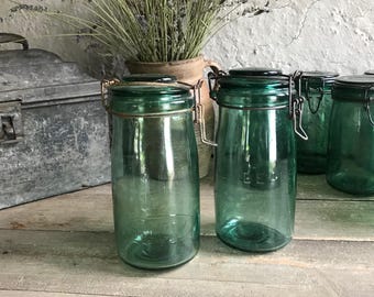 May include: Four vintage green glass jars with metal lids and wire closures. The jars are on a wooden surface.