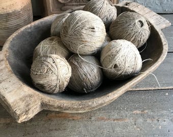May include: A rustic wooden bowl filled with several balls of natural twine. The twine balls vary in size and shade, ranging from light beige to a darker gray-brown. The bowl has a weathered appearance, adding to the vintage aesthetic.