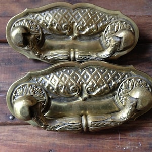 May include: Two ornate brass drawer pulls with a decorative floral design and a diamond pattern. The pulls have a rounded shape and a raised center.