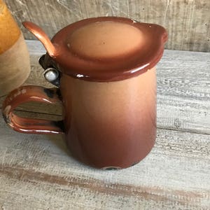 May include: A brown enamelware pitcher with a lid and a handle. The pitcher has a gradient of color from a darker brown to a lighter brown.