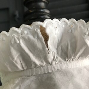 Pu&ograve; includere: Primo piano di un colletto bianco in stile vintage con bordo smerlato. Il colletto presenta delicati dettagli floreali ricamati e occhielli. Il tessuto sembra essere un materiale morbido e leggero.
