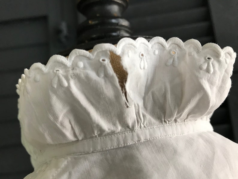 Pu&ograve; includere: Primo piano di un colletto bianco in stile vintage con bordo smerlato. Il colletto presenta delicati dettagli floreali ricamati e occhielli. Il tessuto sembra essere un materiale morbido e leggero.