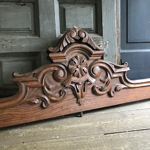 Könnte beinhalten: Ein dunkelbrauner architektonischer Giebel aus Holz mit kunstvollen handgeschnitzten Schnörkeln, Akanthusblattmotiven und einer zentralen Blumenrosette. Das horizontale Stück zeigt ein symmetrisches Design mit Reliefschnitzereien.