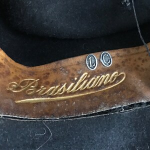 以下が含まれることがあります： 茶色の革製の帽子バンドのクローズアップ。金色の文字で「Brasiliano」と書いてあり、銀色の金属製のタグが2つあり、「L」と「C」と書いてあります。