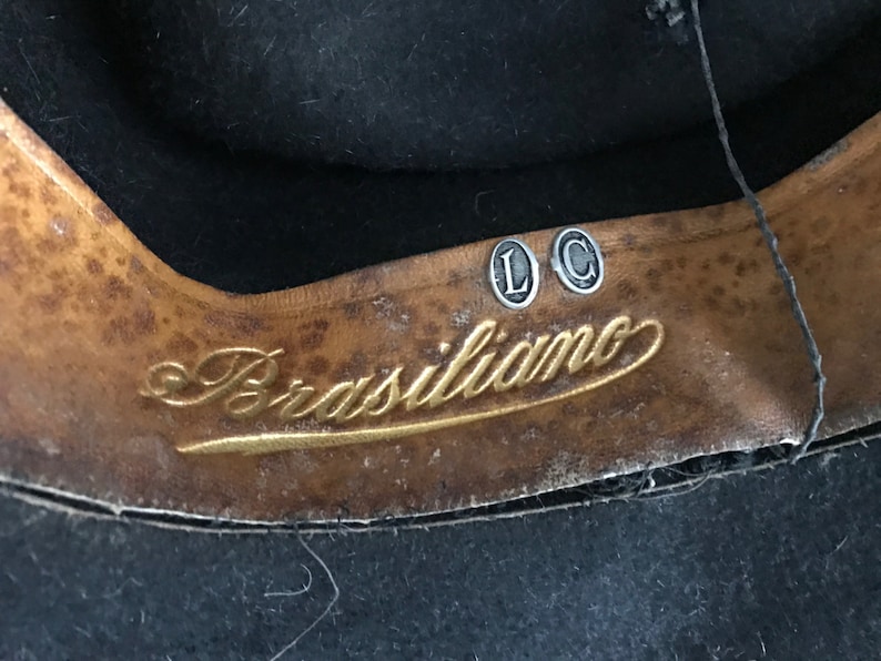 以下が含まれることがあります： 茶色の革製の帽子バンドのクローズアップ。金色の文字で「Brasiliano」と書いてあり、銀色の金属製のタグが2つあり、「L」と「C」と書いてあります。