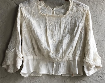 May include: A cream-coloured vintage blouse with detailed floral embroidery. The blouse has a square neckline, three-quarter sleeves, and a gathered waist. It is displayed on a wooden hanger against a textured, neutral background.