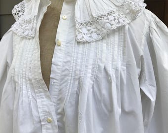 Può includere: Una camicia bianca di cotone con un colletto in pizzo e dettagli plissettati. La camicia ha una chiusura con bottoni sul davanti e maniche lunghe.