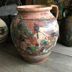 Puede incluir: Una gran jarra de cerámica de terracota redonda con asa. La jarra está decorada con un diseño abstracto colorido en tonos de verde, negro, blanco y rojo.
