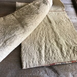May include: A beige linen fabric with a red and orange striped border. The fabric is folded in half and has a rough, textured surface.